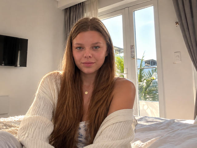 RhonaBells with long brown hair, wearing a white off-shoulder sweater, sitting on a bed, sunlight in a modern room, small gold necklace, cute, slight smile, soft light.