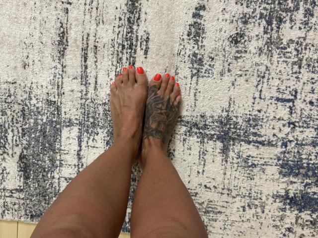 AndraD's feet with red-painted toenails and a large tattoo on her right foot, standing on a textured blue and white rug. Her brown skin is visible.
