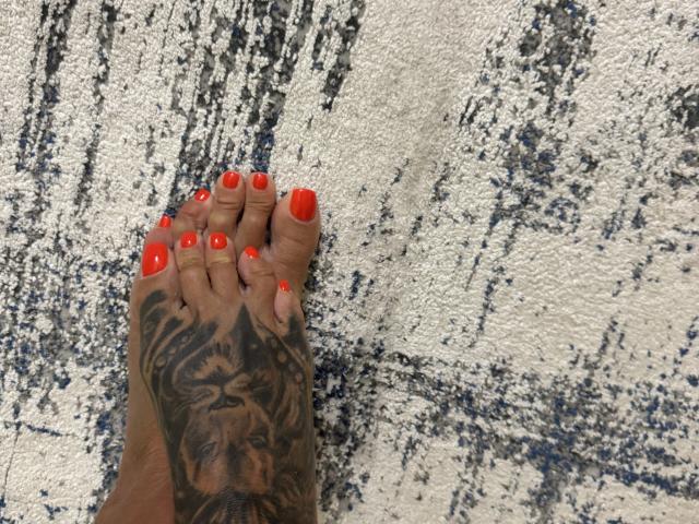 AndraD's foot with bright red toenails, detailed black tiger tattoo on the sole, standing on a textured white and blue carpet.