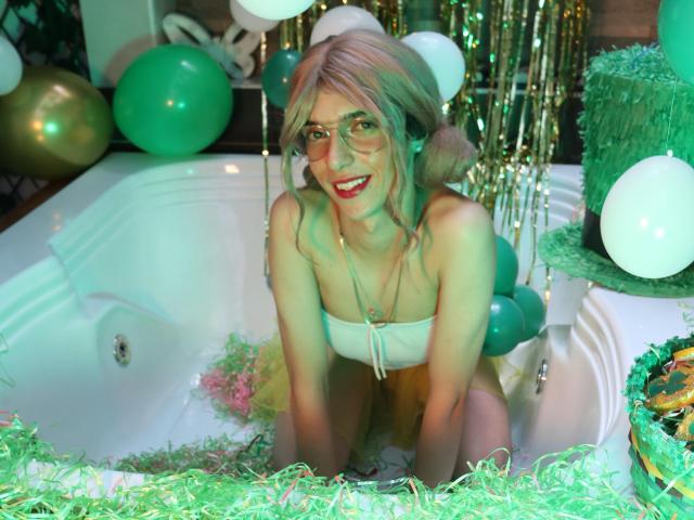 Allison Pearzon, con i capelli rosa, gli occhiali e il rossetto rosso, inginocchiata in una vasca illuminata di verde, indossa un top bianco, ed è circondata da palloncini verdi e bianchi e da festoni verdi..