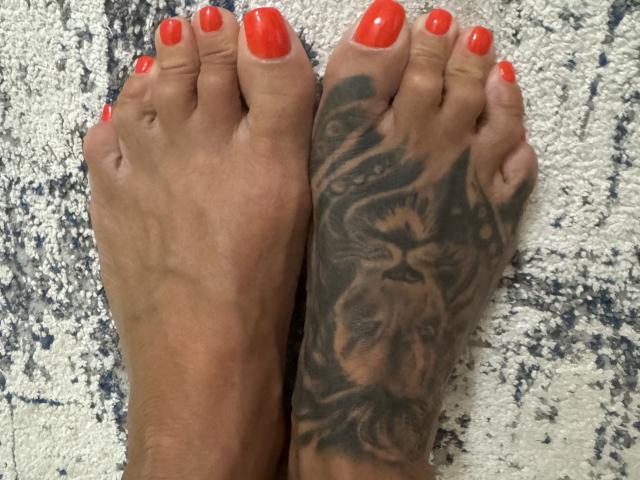 AndraD's feet, with bright red nail polish and a detailed black tiger tattoo on the right sole, standing on a textured carpet.