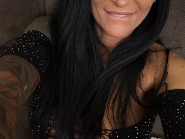 AndraD's smiling face with long black hair, wearing a black sparkly top, showing a tattooed arm and a hint of her small, perky tits.