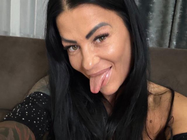 AndraD has long black hair, brown eyes, and is sticking out her tongue. She's wearing a black, sparkly top, with a tattoo on her right arm.