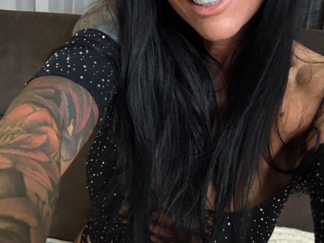 AndraD has long black hair, a tattooed arm with colorful roses, and is wearing a black polka-dot top, showing a slight cleavage and a smile with visible teeth.