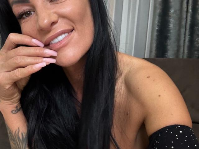 AndraD with long black hair, winking, smiling, showing white teeth, pink nails, tattoo on right hand, small moles on shoulder, wearing black off-shoulder top, brown skin.