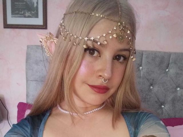 Melani Becker a les cheveux blonds, un piercing au nez et un ornement doré orné de bijoux pendants. Elle porte du rouge à lèvres rose et un collier de perles, et elle regarde l'appareil photo avec un sourire..