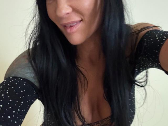 AndraD with long black hair, slightly open mouth, wearing a black, sparkly top with a low neckline, exposing her cleavage and showing a tattoo on her right shoulder.