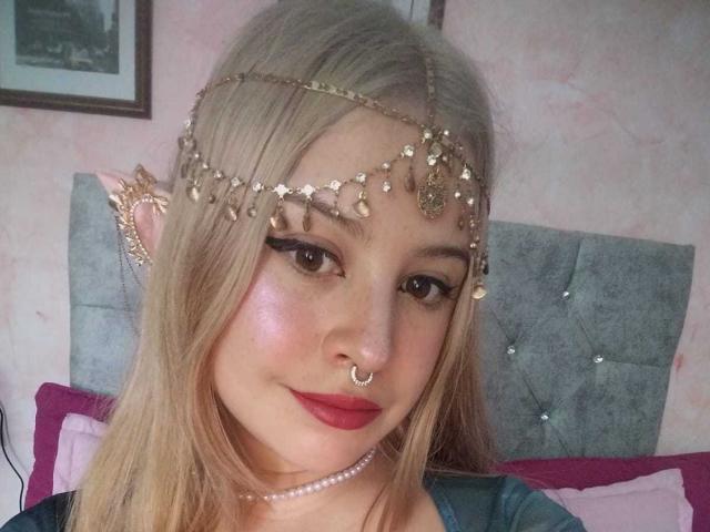 Melani Becker a les cheveux blonds, une chaîne dorée autour du cou, du fard à paupières noir, du blush rose, du rouge à lèvres rouge, un anneau au nez et un collier de perles, et elle est assise sur un lit avec un oreiller violet..