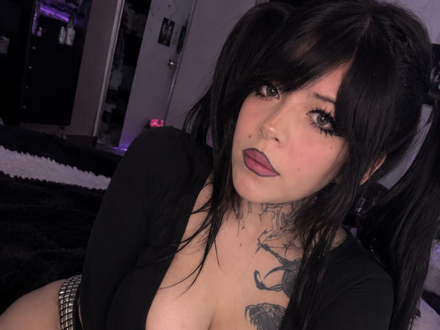 BabeSix with black pigtails, pink lipstick, and tattoos, wearing a low-cut black top, showing her tits, and a studded belt, looking at the camera.