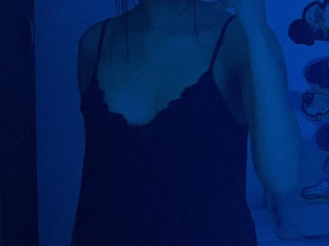 HannaThorne in a black, lace-trimmed, spaghetti-strap nightie, showing her tits under blue, dim lighting, with her arm slightly raised, holding a phone.