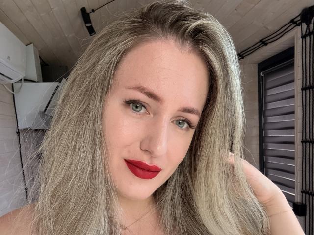 LilaIris has long, messy blonde hair, green eyes, and red lipstick. Her hand is in her hair, and she's smiling softly. She's wearing a necklace.
