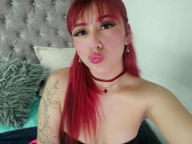 MaddyMills met rood haar, volle roze lippen, dikke wimpers, een tatoeage op de linkerarm, een choker ketting, kleine oorbellen, een pluizig grijs kussen en een gestoffeerd grijs hoofdeinde op de achtergrond..