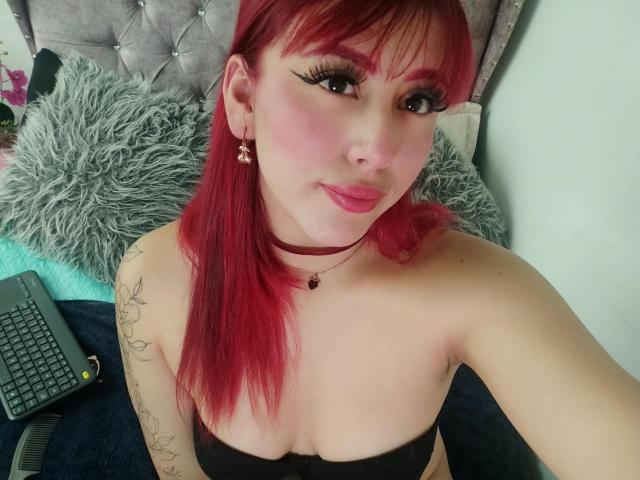 MaddyMills met rood haar, een zwarte top zonder bandjes, waarbij haar borsten zichtbaar zijn, roze lippenstift, zwarte eyeliner, een choker, een tatoeage op haar arm, een laptop op het bed, een grijze hoofdbord en een pluizig kussen..