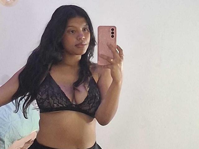 PauletteDelacroix with long black hair, wearing a black lace bra that shows her big tits, holding a pink phone, taking a mirror selfie, looking slightly to the left.