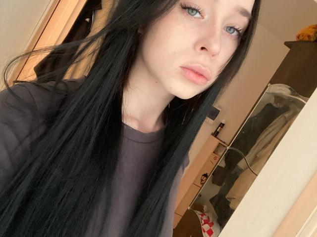 MurKristian a de longs cheveux noirs, une peau pâle, des yeux bleus et des lèvres roses. Elle porte une chemise sombre et regarde directement l'appareil photo avec une expression légèrement boudeuse..