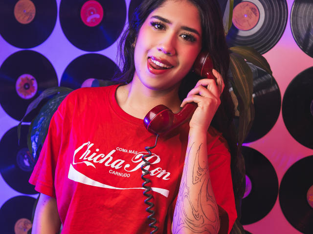 HannaThorne with long dark hair, red Coca-Cola shirt, holding red corded phone, tattooed arm, smiling, pink and purple lighting, vinyl records in background.