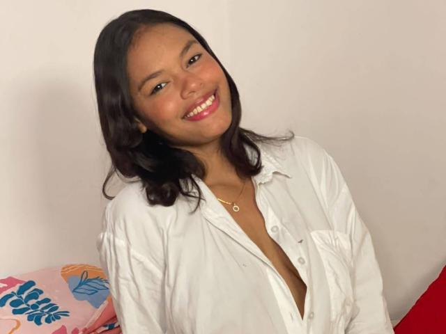 AmbarQueent, avec ses cheveux bruns foncés, souriante, portant une chemise blanche ouverte qui dévoile sa poitrine, et un collier en or, assise contre un mur blanc..