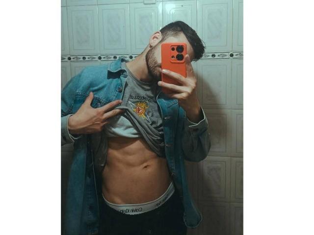 MaddyAndLogan's toned abs, visible under a raised denim shirt and gray tee, with a hand on chest. He's holding an orange phone, wearing black underwear.