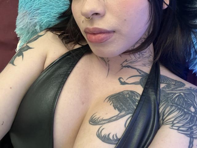 BabeSix with pouty lips, nose ring, and tattoos, wearing a black leather halter, showing large tits and detailed bird tattoos on her chest and shoulder.