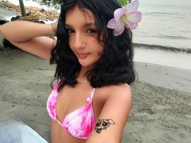 PaulineRey with medium tits in a pink tie-dye bikini, black wavy hair, pink plumeria in hair, tattoo on right arm, standing on sandy beach, smiling.