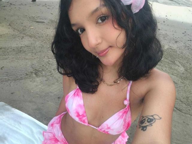 PaulineRey with wavy black hair, pink tie-dye bikini, small tits, tattoo on right arm, cute smile, pink headband, sitting on sandy beach, white towel in background.