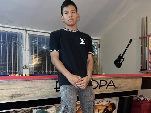 RoomieSsecretsss stands in front of a pool table, wearing a black "V" shirt and gray distressed jeans, with hands crossed, short black hair, and a serious expression.