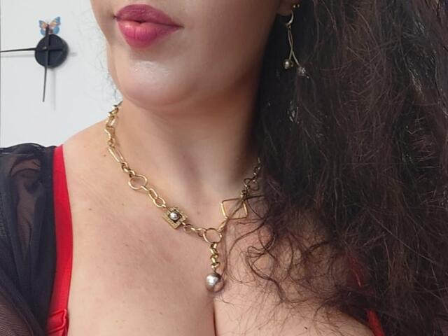 VanessaSquirtt's face with red lipstick, gold chain necklace, and earrings, showing her cleavage in a red top with black mesh. Her curly brown hair is on the right.