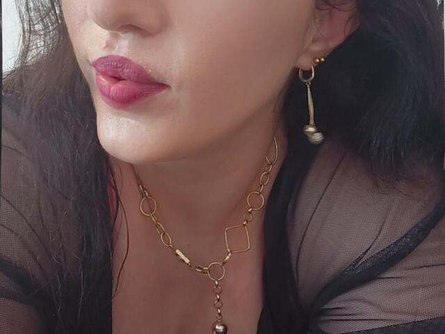 VanessaSquirtt's face, showing glossy pink lips, a gold chain necklace, and black sheer top, with a gold hoop earring in her right ear, and wet skin.