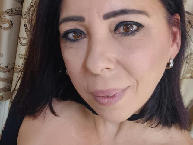 LauraSin has brown eyes, dark brown hair, and is wearing black eyeliner and pink lipstick. She's smiling with slightly parted pink lips, wearing small diamond earrings.