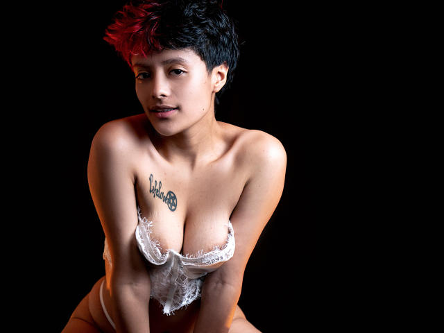 KvsKael with short black hair, red highlights, tan skin, small tattoos, wearing white lace, exposing her tits, kneeling, black background, looking at camera, slightly smiling.