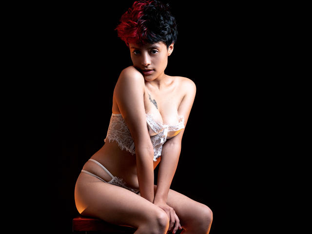 KvsKael con capelli corti neri e rossi, indossa un reggiseno e una biancheria intima di pizzo bianchi, seduta su uno sgabello rosso, mani sulle ginocchia, guarda la fotocamera, sfondo scuro..