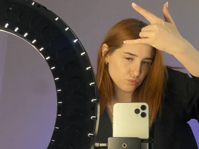 VeolaVanhoecke, con i capelli rossi, fa un gesto di pace, tenendo un telefono con una luce ad anello di fronte a un grande pneumatico nero in un selfie..