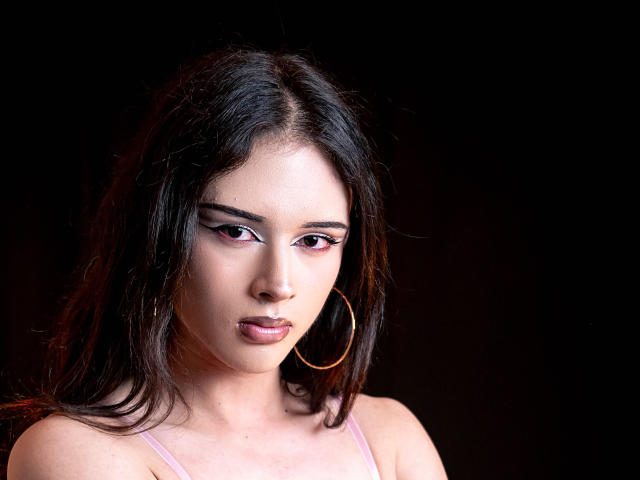 KvsKael ha i capelli scuri e ondulati, il trucco occhi rosa e grandi orecchini a cerchio. Guarda leggermente verso il basso con le labbra leggermente aperte, mostrando il suo naso piccolo e rotondo e la bocca leggermente rivolta verso l'alto..