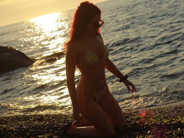 Sunset beach, AlexXas kneeling, wearing a yellow bikini, tits in a triangular top, small butt, red hair, sunglasses, water reflecting sunlight, pebbled shore, right hand touching water.
