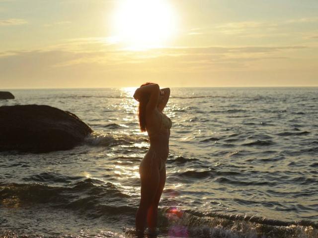 Silhouetted AlexXas stands in shallow ocean water, hands behind head, sunlight reflecting off wet skin, showing small tits and slim ass, with rocky shore to the left.