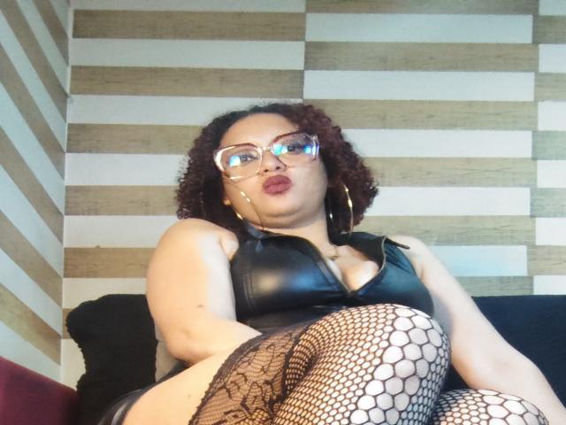 VioletaSwitch mit lockigem, rotem Haar, Brille, einem schwarzen Lederoberteil und Fischernetzstrümpfen, die die Lippen zu einem Schmollmund formt, sitzt auf einer schwarzen Couch und hält ein Smartphone für ein Selfie..