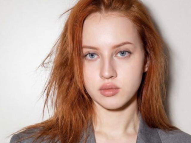 JannaCorsaro has pale skin, red messy hair, blue eyes, and pouty pink lips. She's wearing a gray shirt, and her tits are small and barely visible.