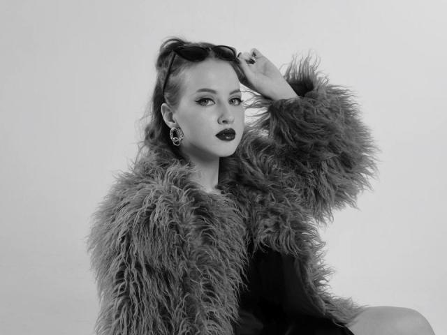 JannaCorsaro with fluffy coat, dark lipstick, and earrings, holding sunglasses on her head, showing off her tits, looking confident and sexy. Black and white, high-contrast photo.