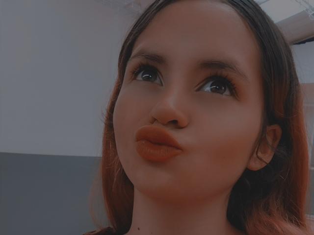LesleyGray with pouty, orange lipstick, looking up with big brown eyes, dark brown hair, and a slightly flushed face. Her ass is not visible.