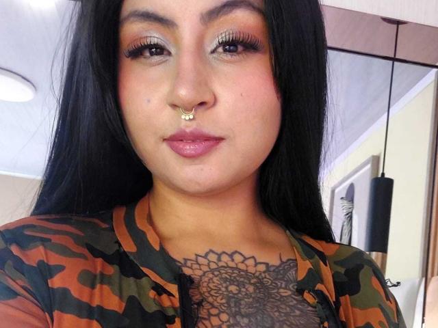 AngelaJoness tiene el pelo largo y negro, ojos marrones, un piercing en la nariz y una camisa de camuflaje. Está mostrando un tatuaje detallado de un mandala en su pecho, con unos labios rosados y brillantes..