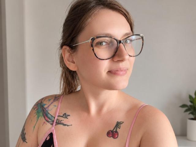 AggieSexy trägt eine Brille, hat braunes Haar und Tätowierungen an Schulter und Brust. Sie lächelt und trägt ein pinkes Top mit Spaghetti-Trägern, das ihre Brüste zeigt und eine Tätowierung in Form einer Kirsche..