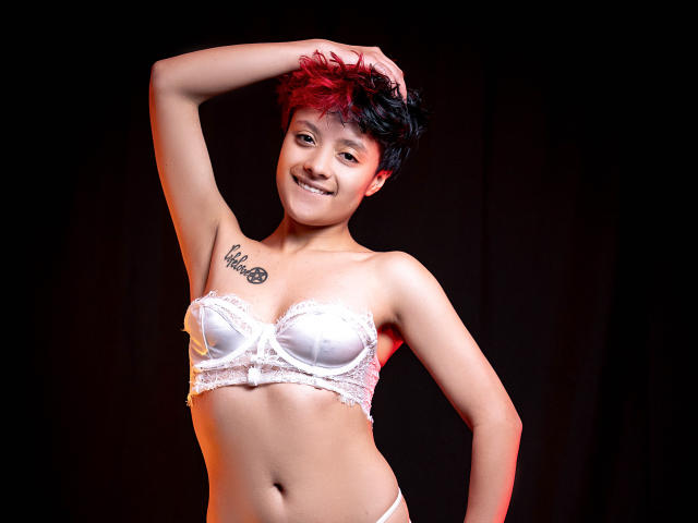 KvsKael with short black and red hair, smiling, wearing white lace bra, hand on head, tattoo on left chest, standing against black background, cute, small tits, slim.