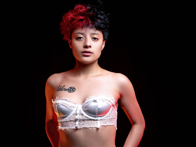 KvsKael, con capelli ricci corti, rossi e neri, indossa un reggiseno bianco senza spalline in pizzo, mostrando piccoli seni, con un tatuaggio sul petto sinistro, in piedi su uno sfondo scuro..