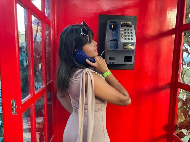 AshlyLee with brown hair, wearing a beige dress, talking on a blue phone in a red phone booth, has sunglasses on her head and a green wristband.