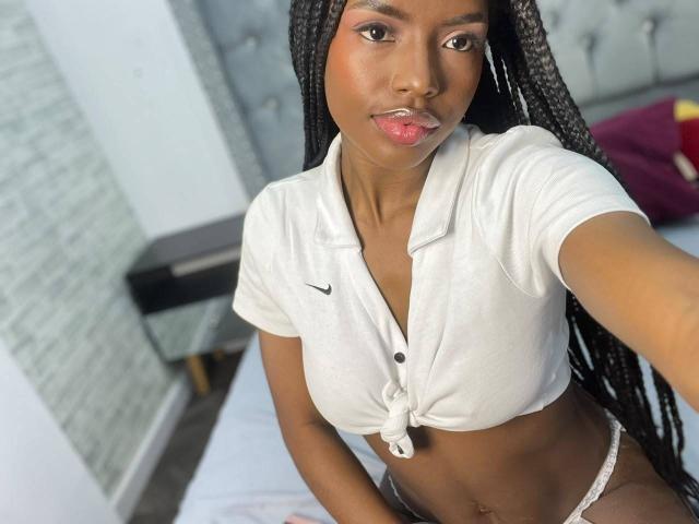 EllysRose, met donkere huid, lange vlechten, roze lippen, draagt een witte polo met knoopjes, waarbij haar borsten zichtbaar zijn, en witte kantonderbroek, en maakt een selfie..