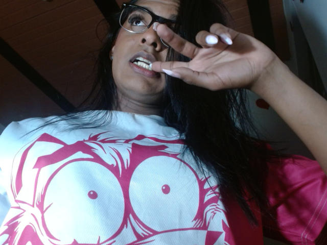 IdiiaLatina with dark hair, glasses, and white shirt with red pattern, biting nail, showing her large tits, looking down at camera. She's wearing a pink pillow in background.