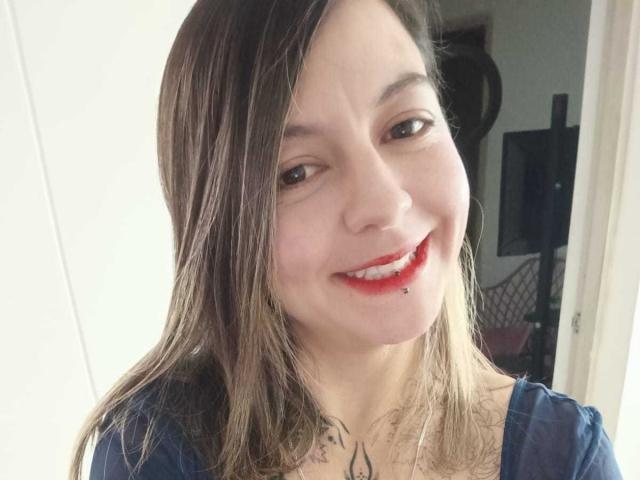 ArianaBlonde with light brown hair, red lipstick, and a lip piercing, smiling at the camera, wearing a blue shirt, showing her tits slightly, in a selfie.