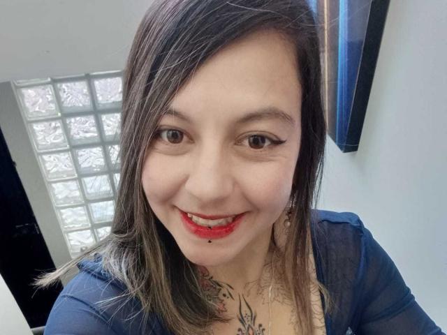 ArianaBlonde with a cute smile, red lipstick, and black eyeliner, has a small tattoo on her chest, brown hair, and is wearing a sheer blue top.