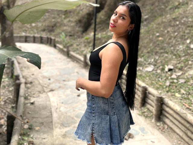 MelodyRoss with long black hair, red lipstick, black tank top, and blue denim skirt, standing on a stone path, looking over her shoulder, showing her ass.