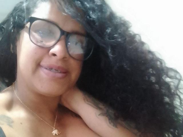 ZoehFerraro, con cabello negro rizado, gafas y un collar dorado, sonríe con un brazo cruzado sobre su pecho desnudo, mostrando pequeños tatuajes. Sus pechos son parcialmente visibles..